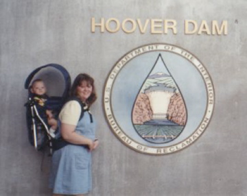 Woman and child pose beside the Hoover Dam sign. A woman in a blue dress holds a baby in a carrier next to a Hoover Dam sign with a water drop emblem.