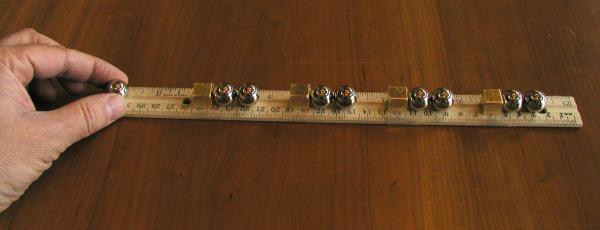 A hand places small metallic cylinders along a ruler on a wooden table. A hand holds a ruler with multiple metallic cylindrical objects placed along its length on a wooden surface.