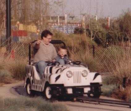 A man and child enjoy a ride in a zebra-themed go-kart. A man and a young child ride in a white zebra-patterned go-kart on a track.