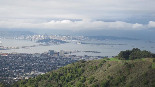 View from Hill Road over San Francisco Bay. A distant view of a city skyline across a bay, with hills in the foreground and clouds overhead.