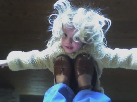 A young child with curly blonde hair wearing a white fuzzy jacket and brown shoes, sitting with arms outstretched.