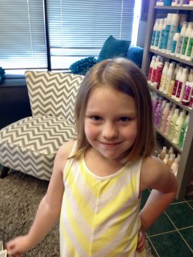 A young girl with shoulder-length brown hair wearing a yellow and white striped tank top stands in front of shelves filled with bottles, smiling at the camera.