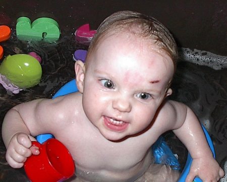 A baby in a bathtub holding a red cup with colorful bath toys nearby.