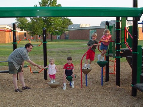 A man and four children playing on a playground structure with colorful equipment.