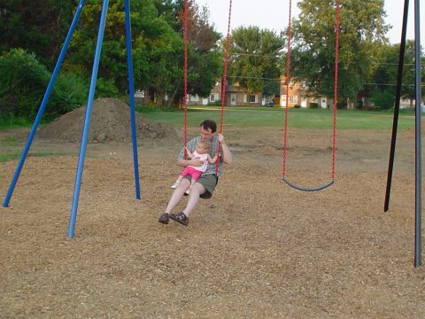 A man holding a small child on a swing at a playground with wood chips and trees in the background.
