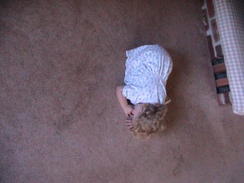 A young child with curly hair wearing a white patterned dress lies on their side on a brown carpet next to a brick structure.