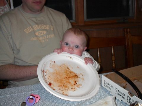 A baby holds a dirty white paper plate in front of its face while sitting at a table with pizza box and pacifier nearby.