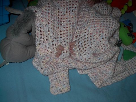 A baby's feet peek out from under a pink crocheted blanket with sparkles, surrounded by stuffed animals on a blue surface.