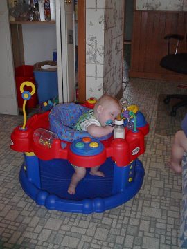 A baby in a colorful activity walker sitting on a tiled floor.