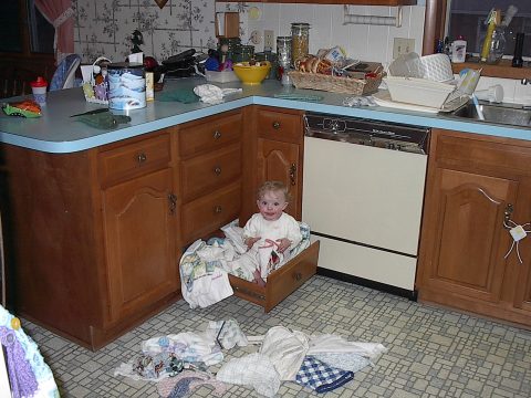 A young child sits inside an open kitchen drawer filled with baby clothes, surrounded by scattered laundry on the floor in a cluttered home kitchen.