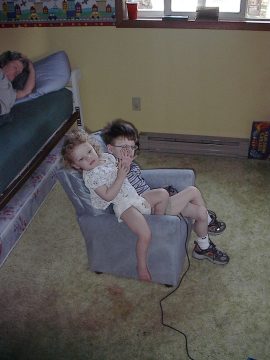 Two young children sitting on a small blue armchair in a bedroom with a bed and window.