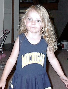A young girl with curly blonde hair wearing a navy blue Michigan dress stands indoors.