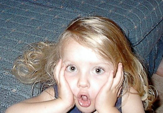 A young child with blonde hair holding their face in a surprised expression.