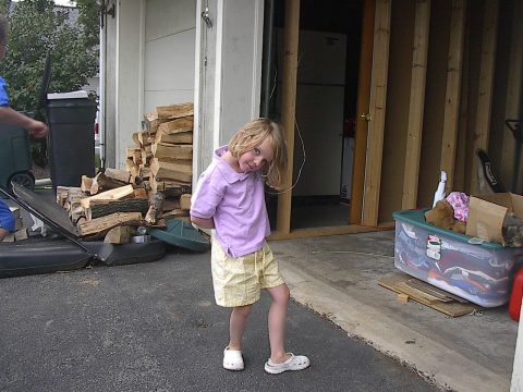A young girl in a purple shirt and yellow shorts stands outside a garage with wood stacked nearby.