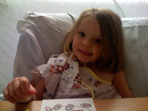 A young girl with light brown hair wearing a patterned top smiles while sitting in a hospital bed.
