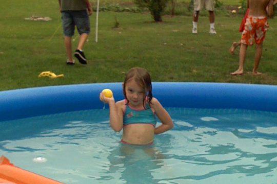 A young girl in a blue swimsuit stands waist-deep in an inflatable pool holding a yellow ball.