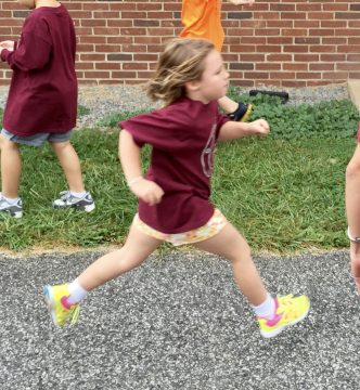 A young girl in a maroon shirt and colorful shorts runs across asphalt while other children stand nearby on grass.