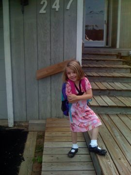 A young girl in a pink floral dress stands on a wooden porch with a backpack.