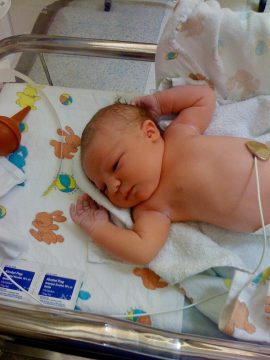 A newborn baby sleeping in a hospital bassinet with medical monitoring equipment attached.