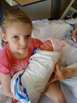 A young girl in a pink shirt holds a swaddled newborn baby wearing a pink hat in what appears to be a hospital room.
