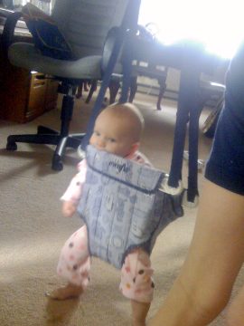 A baby in a walking harness standing on carpet near an office chair.