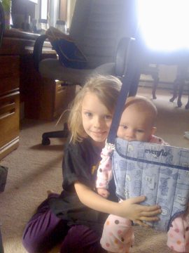 Two young children sitting on a carpeted floor indoors; one holds a blue patterned baby seat with visible text.