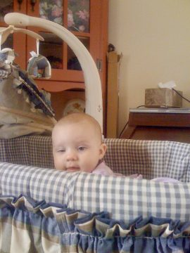 A baby with short hair looking forward from inside a blue and white checkered crib.