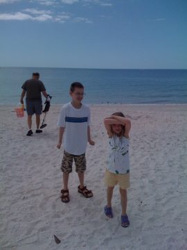 Two children standing on a sandy beach with the ocean in the background; one child holds hands over eyes.