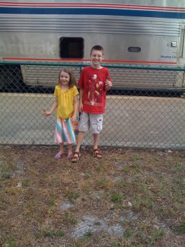 Two children standing in front of a train with a fence between them.