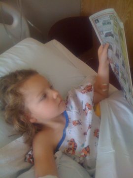 A young girl with curly hair lies in a hospital bed wearing tiger-patterned pajamas and reading a newspaper while looking up.