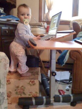 A toddler with a pacifier stands on a floral box reaching for a laptop on a raised desk.