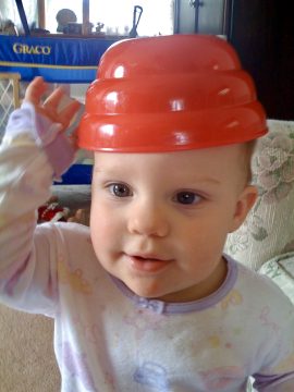 A baby wearing a red plastic bowl on their head, looking at the camera with one hand raised.