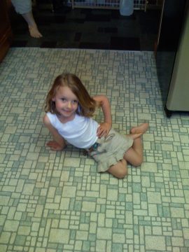 A young girl with light brown hair wearing a white tank top and beige shorts sits on a patterned floor looking up at the camera.