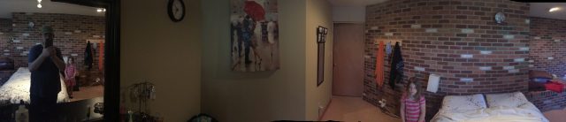 A panoramic view of a bedroom with brick walls, a bed, a girl standing near the wall, and a person taking a photo in a mirror.
