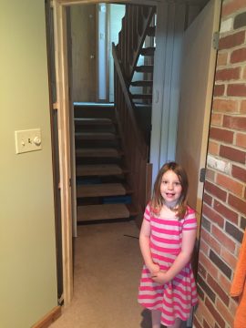 A young girl in a pink and white striped dress stands smiling near a doorway with stairs leading upward.
