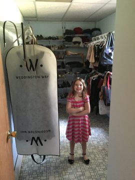 A young girl in a pink striped dress stands with arms crossed next to a large garment bag hanging in a closet filled with shoes and clothes.