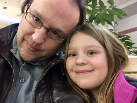 A man wearing glasses and a leather jacket smiles closely with a young girl in a pink jacket; they are indoors near green plants.
