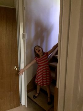 A young girl in a red and white striped dress stands on stairs with arms outstretched near a door.