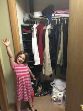 A young girl in a pink striped dress stands smiling with one arm raised inside a cluttered closet filled with hanging coats, shelves, and household items.