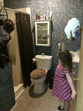 A young girl in a pink striped dress stands in a bathroom with blue patterned wallpaper, reaching toward a mirror while holding a small object.