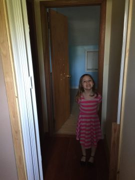 A young girl in a pink and white striped dress stands smiling in a doorway.