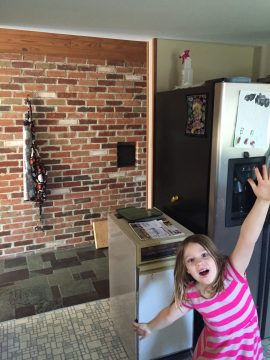 A young girl in a pink striped dress stands excitedly in a kitchen with exposed brick walls and stainless steel appliances.