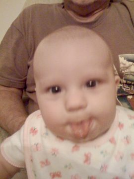 A baby with a patterned bib sticking its tongue out while sitting in front of an adult wearing a brown shirt.
