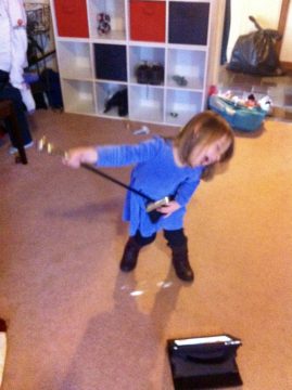 A young child in a blue dress holds a long black stick with a small object at the end, standing on carpet near a storage unit.