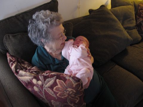 An elderly woman with gray curly hair and glasses holds a sleeping newborn baby wrapped in pink clothing on a dark couch.