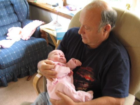 An older man in a dark t-shirt sits in a beige armchair holding a sleeping newborn baby wrapped in pink clothing.