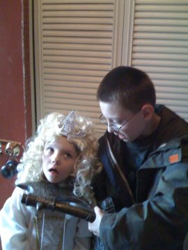 A person in a blonde wig and tiara looks surprised while another person holds a large prop object near them.
