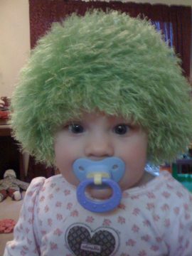 A baby wearing a green fuzzy wig and a blue pacifier with a heart design on the shirt.