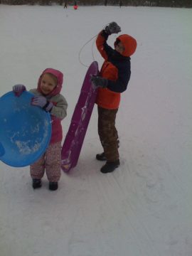 Two children in winter clothing holding sleds on a snowy field.
