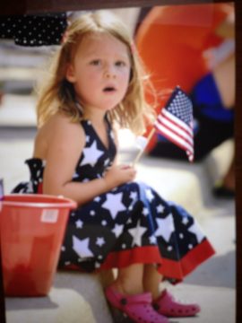 A young girl in a patriotic dress holding a small American flag.